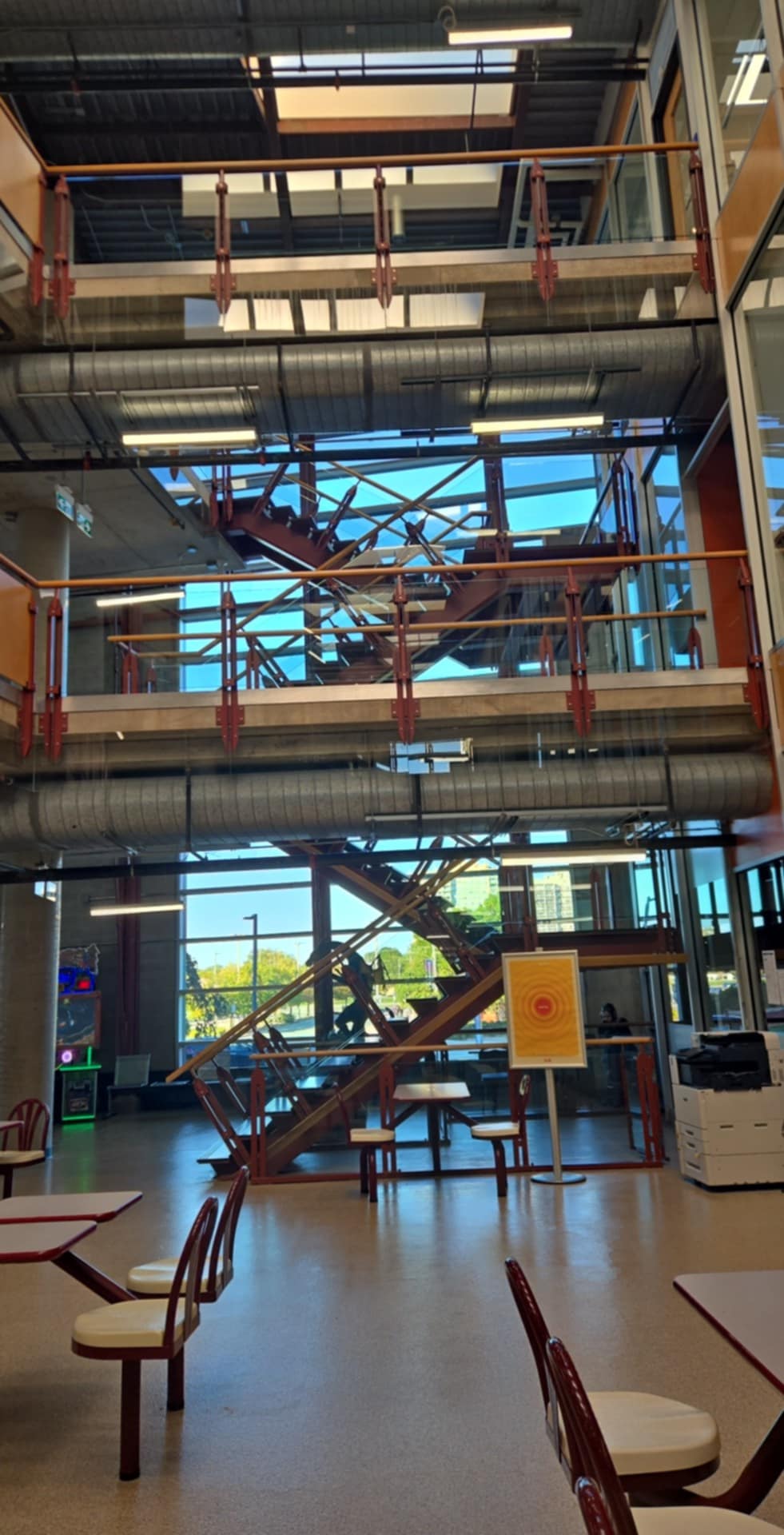 This is the interior of the S wing building, showcasing the architectural structure of the staircase. This is a busy staircase during the school hours. 
      The exposed steel, duct and staircase is a type of industrial style, most common style in office, big buildings/institution. This space is also a social hub, 
      as provided with table and chairs. In terms of colour, the windows provide natural sunlight and gives people the feeling of alertness. 